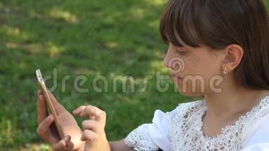 美丽的女孩使用智能手机正在一个美丽的绿色公园的长凳上写一封信。 慢动作。 青年